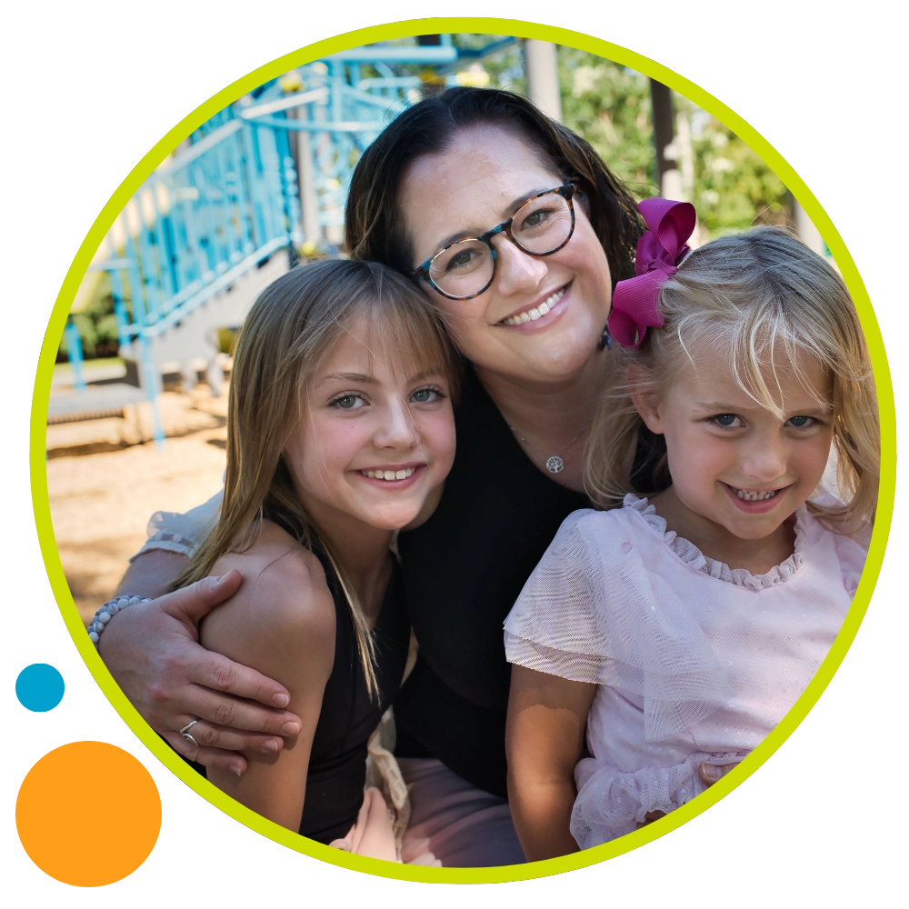 A woman with glasses smiles and hugs two young girls, all sitting outside near playground equipment. The girls are smiling; one has a pink bow in her hair. The photo is framed with a yellow-green circular border. A woman with glasses smiles and hugs two young girls, all sitting outside near playground equipment. The girls are smiling; one has a pink bow in her hair. The photo is framed with a yellow-green circular border.