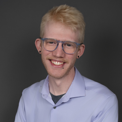 A young person with short blond hair, light skin, and blue glasses smiles at the camera. They are wearing a light purple collared shirt over a gray t-shirt, with a dark gray background behind them.