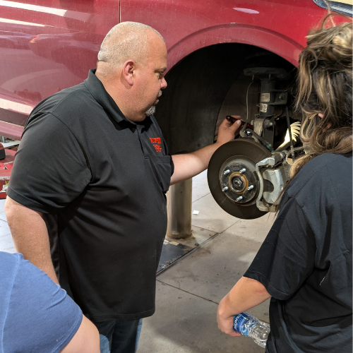 A man in a black polo shirt explains car brake components to two people near a lifted red vehicle in a garage. One person holds a water bottle, while the man points to parts inside the wheel well.