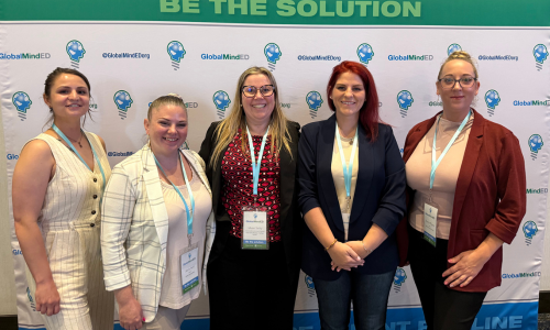 Five women stand side by side, smiling at the camera, in front of a backdrop with the GlobalMindED logo and the phrase "BE THE SOLUTION" at the top. All are wearing conference badges.