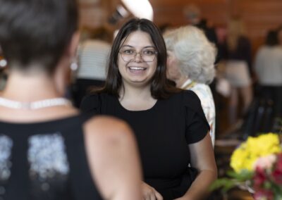 A young woman with long brown hair and glasses, wearing a black dress, smiles while talking to another person at an indoor event. People and flowers are visible in the blurred background.