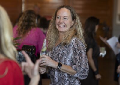 A woman with wavy blonde hair, wearing a snake-print blouse and a watch, smiles while holding a drink at a social gathering. Several people are visible in the background, some blurred.