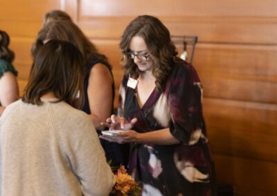 A woman in a patterned dress and glasses looks down at a tablet or phone while talking to another woman in a light sweater, with others and wood-paneled walls in the background.