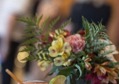A pink cocktail with a lime wedge sits on a table beside a bouquet of colorful flowers and a "Scan to Donate" sign with a QR code; blurred people are visible in the background.