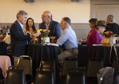 A group of people in business attire converse around tall tables with flower centerpieces at a social or networking event. Some are seated, others stand, and drinks are visible on the tables.