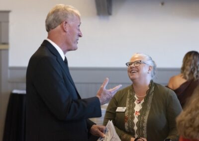 A man in a suit and a woman with glasses and gray hair, wearing a green jacket, are smiling and talking at an indoor event. The woman holds a piece of paper, and other people are blurred in the background.