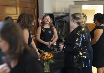 A group of women dressed in formal attire gathers and converses indoors near a table with a flower arrangement and a black box. One woman in a black dress stands smiling among them. The scene appears to be a social event.