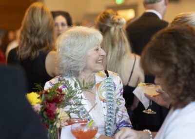 An older woman with gray hair smiles while holding a drink at a social event, surrounded by other people. She wears a colorful jacket and there are flowers on the table in front of her. The atmosphere is bright and lively.