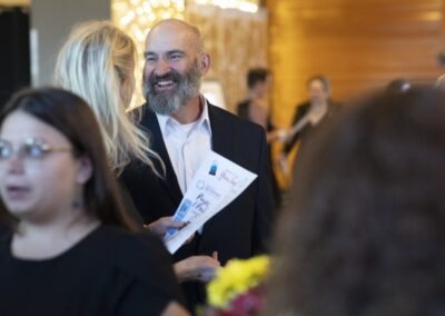 A man in a suit smiles and chats while holding a program at a lively indoor event, surrounded by people and warm lighting, with some blurred figures in the foreground.