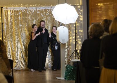 Three people in formal attire pose and smile for a photo at an event, standing in front of a gold sequin backdrop with string lights. A photo booth with a white umbrella light is set up in front of them.