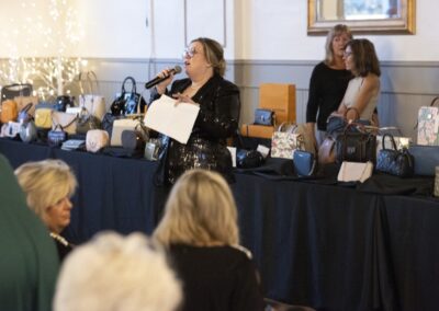 A woman in a black sequin jacket speaks into a microphone, holding papers, in front of a table displaying various handbags. Several women are seated and standing around her in a well-lit room.