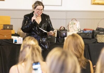A woman in a black sequin blazer speaks into a microphone and holds papers, standing in front of a table with designer handbags and gift bags as an audience looks on.