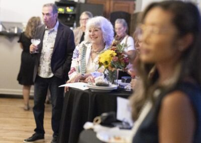 People stand and sit around high tables at a social event. A woman with white hair and a floral jacket smiles near a flower centerpiece, while others mingle and chat in a warmly lit room.