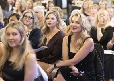 A group of women sit closely together at an indoor event, attentively watching something off-camera. Most women are dressed formally and appear engaged, with some smiling and others looking serious.