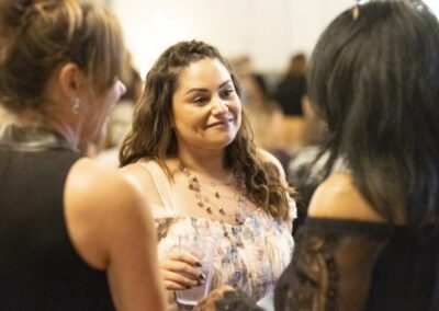 Three women are standing together indoors, engaged in conversation. One woman in the center, holding a drink, is smiling warmly at the others. The background is softly blurred, highlighting their interaction.
