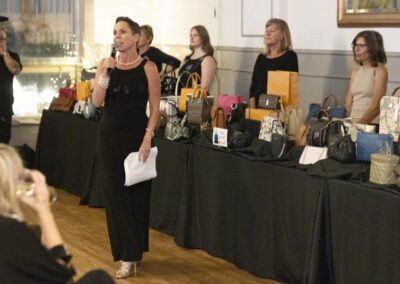 A woman in a black dress speaks into a microphone at an indoor event, standing in front of tables displaying various handbags and boxed items, with several women and an audience in the background.