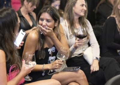 Two women sit closely together at an event, laughing while holding wine glasses. One checks her phone, and people in the background also appear to be enjoying themselves with drinks.