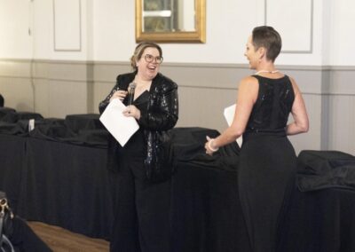 Two women dressed in black evening attire stand in front of a long table covered with a black cloth. One woman holds a microphone and papers, smiling at the other woman, who is also smiling.