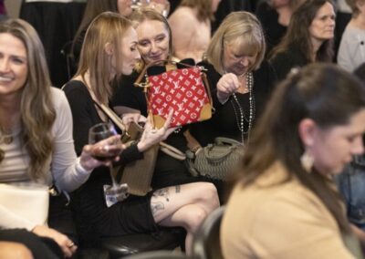 A group of women sitting closely together at an indoor event, some smiling and talking. One woman holds up a red designer handbag, and another looks into her own purse. The atmosphere appears lively and social.