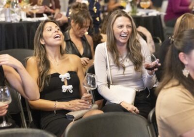 Two women sit side by side at an event, laughing and holding wine glasses. Other people are seated around them at tables with drinks, and the atmosphere appears lively and joyful.