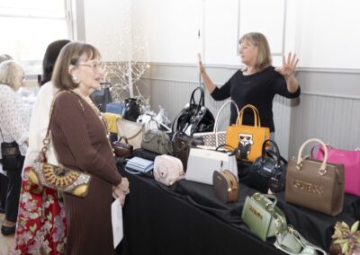 Two women stand at a table displaying various handbags, while another woman stands behind the table gesturing with her hands. Several colorful purses are arranged on the table in front of them.