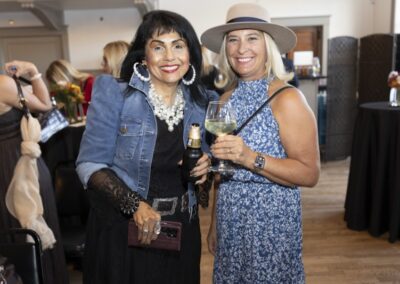 Two women smiling at an indoor event, one wearing a denim jacket and large earrings holding a drink bottle, the other in a blue floral dress and wide-brimmed hat holding a glass of white wine. People and tables are in the background.