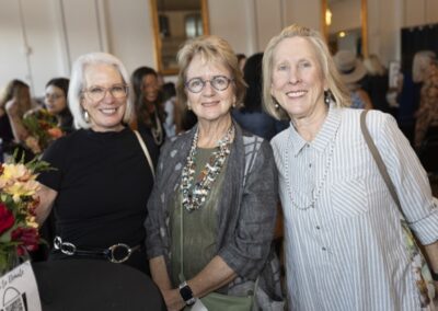 Three smiling women with light hair and glasses stand together at an indoor event. They are dressed in casual, stylish clothing and are surrounded by other people. A bouquet of flowers is visible on a table nearby.