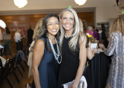Two women smiling and posing together at an indoor event, dressed in elegant attire. People are mingling in the background near tables, under warm lighting.