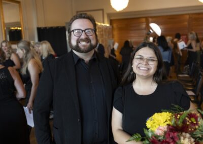 A smiling man and woman, both wearing black outfits, stand together indoors. The woman holds a bouquet of flowers, and people are gathered in the background, suggesting a celebratory event.