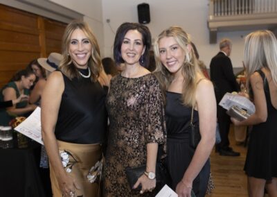 Three women dressed in elegant attire smile together at an indoor event. Other attendees can be seen in the background, some holding programs in a warmly lit room with wooden walls and floors.