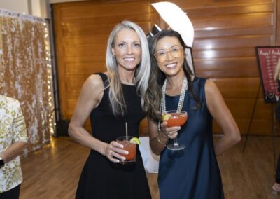 Two women dressed in elegant black dresses smile at the camera while holding cocktails. The background features wood paneling, string lights, and a sequined curtain, creating a festive atmosphere.