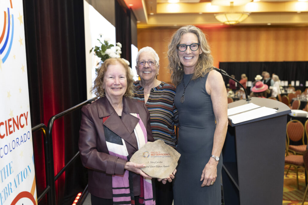 Three smiling women stand together at an indoor event. The woman on the left holds a stone-shaped award, while the other two stand beside her near a podium. Round tables and people are visible in the background.